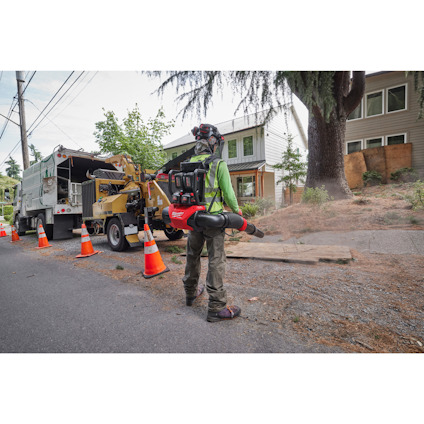 Person använder en ryggsäckslövblåsare på en gångväg nära en lastbil och en flismaskin. Trafikkoner är utplacerade.