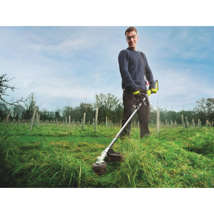 Person anväder en grästrimmerav märket Ryobi på en gräsmatta med trädplantage i bakgrunden.
