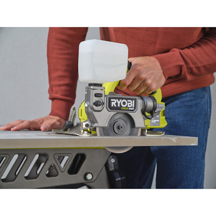Person använder grön och grå kakelsåg med text "RYOBI ONE+" för att skära en platta på arbetsbänk.