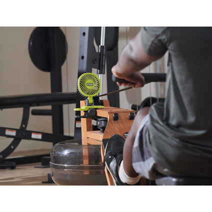 Person tränar på en roddmaskin i ett gym, med en liten fläkt med klämfäste av märket RYOBI  monterad på trästrukturen.