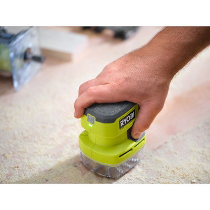 En hand håller i en grön bordsdammsugare med märket "RYOBI" på ett träbord.