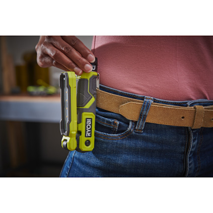 Hand håller en grön och svart Inspektionslampa av märket RYOBI  vid bältet på en persons jeans.
