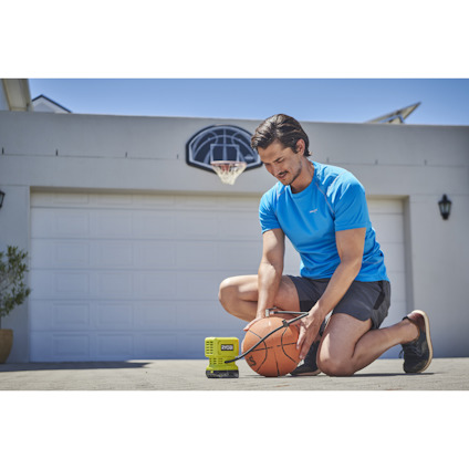 Man i blå t-shirt pumpar en basketboll med en miniluftpump av märket Ryobi nära en garageport, basketkorg syns i bakgrunden.