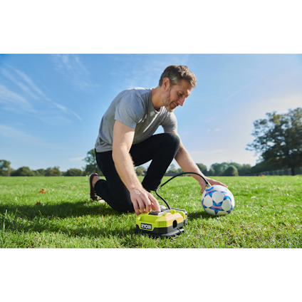 Man använder en grön multikompressor av märket Ryobi för att fylla en fotboll på en gräsmatta.