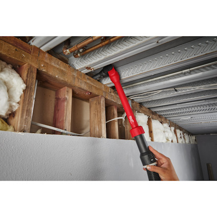 Hand holding a red tool accessing pipes above an unfinished ceiling with exposed insulation and wooden beams.