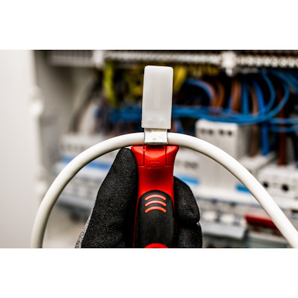 Hand i skyddshandske håller en kabelskaltång nära en vit kabel, suddig bakgrund med kablar.