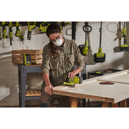 Person slipar en träskiva med en batteridriven excenterslip av märket RYOBI, omgiven av verktyg på en vägg och en hylla.