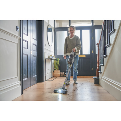 En person dammsuger en hall med trägolv med en skaftdammsugare av märket Ryobi Det finns en spegel och växter i bakgrunden.