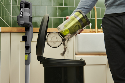 En person tömmer en dammsugarbehållare i en soptunna. Skaftdammsugaren av märket RYOBI står mot en köksbänk.