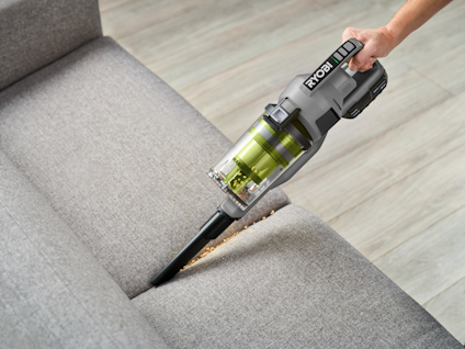 Hand håller en Ryobi skaftdammsugare som suger damm från en grå soffa.