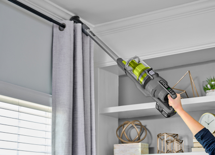 Person som använder en grå RYOBI skaftdammsugare för att rengöra gardinstång nära taket.