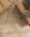Ljusa träplankor i fiskbensmönster, kuddar i brunt tyg och en grön filt delvis synliga.