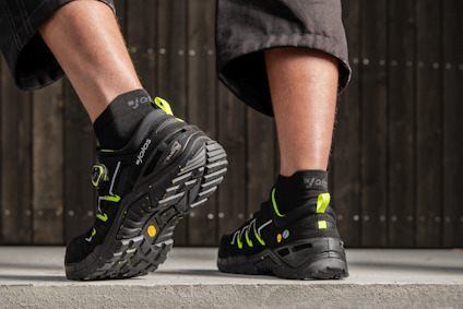 Två fötter i svarta och gröna säkerhetsskor, set bakifrån. Skorna har grova sulor och krokodilmönstrad stabilitet.