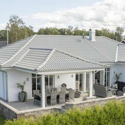 En vit villa med grått tak, stor uteplats med utemöbler och omgivande grön växtlighet.
