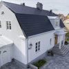 Vit villa med svart tegeltak, kupor och trädörr. Lanternor på fasaden. Grå marksten framför huset.