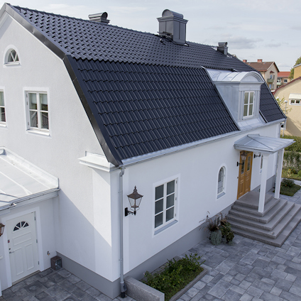 Vit villa med svart tegeltak, kupor och trädörr. Lanternor på fasaden. Grå marksten framför huset.