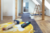 Pensel och måttverktyg på en gul hink i ett rum under renovering; stege i bakgrunden.