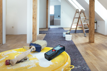 Pensel och måttverktyg på en gul hink i ett rum under renovering; stege i bakgrunden.
