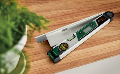 Digital vinkelmätare i metall och plast med inbyggt vattenpass på ett träbord, växt i förgrunden.
