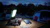 En bärbar lampa och powerbank lyser upp ett bord utomhus bredvid en paviljong och en laddande smartphone.