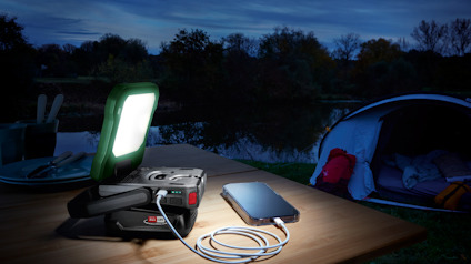 En bärbar lampa och powerbank lyser upp ett bord utomhus bredvid en paviljong och en laddande smartphone.