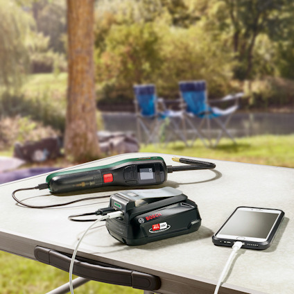 Borrmaskin och smartphone laddas med ett batteripaket på ett campingbord vid en sjö.