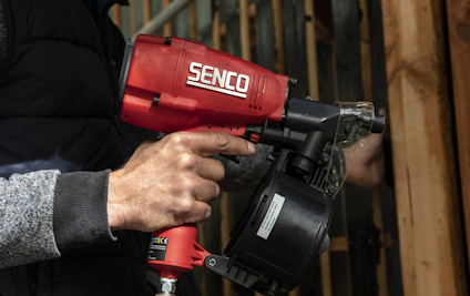 Person som håller en röd spikpistol med märket "Senco", fäster material mot trästödmur.