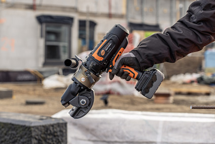 Hand håller i ett orange och svart industriellt verktyg med batteri, bakgrund av byggarbetsplats.