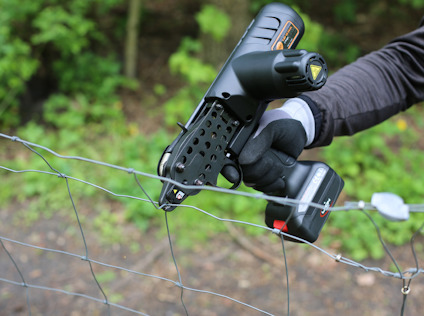 En person håller i en elektrisk verktygspistol, intill ett metallstängsel, omgiven av grönska.