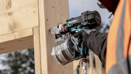 En person i arbetskläder använder en Makita spikpistol på en träkonstruktion.