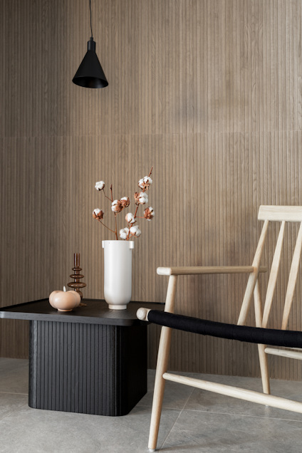 Trästol och bord med svart skiva, vas med bomullsblommor, hängande svart lampa, beige ribbad vägg.