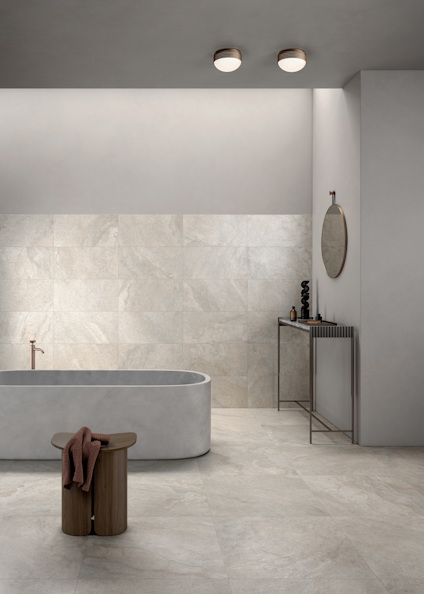 Modernt badrum med grått badkar, rund spegel, brun pall och grå väggar och golv.