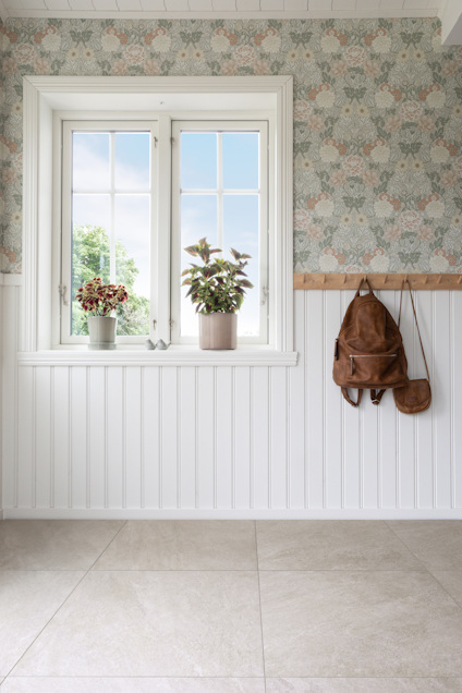 Fönster med krukväxter på fönsterbänk, blommig tapet och väggpanel, ryggsäckar hängande på väggen.