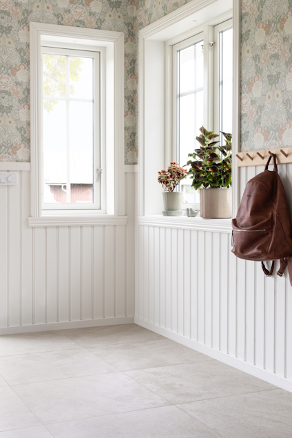 Ljust rum med blommigt tapetmönster, vita paneler, fönster, växter i krukor och en brun läderväska på väggen.