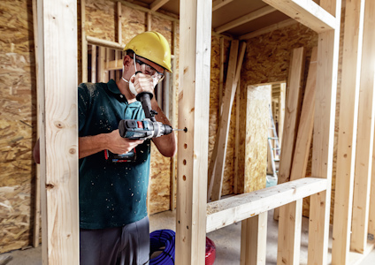 Person som använder en borrmaskin på en byggarbetsplats, omgiven av träreglar och byggmaterial.