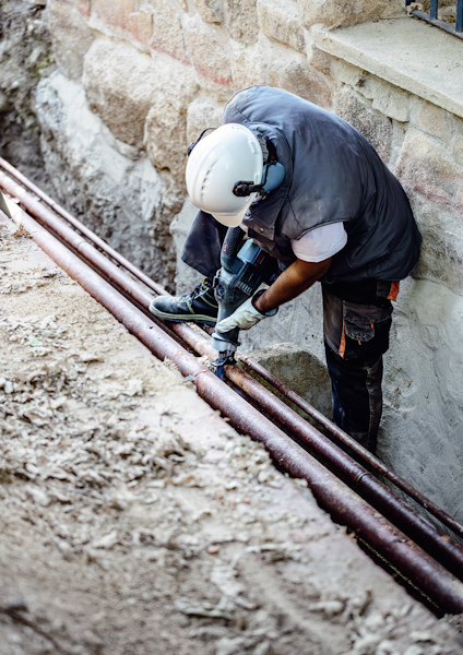 Byggarbetare med hjälm arbetar med rör i ett dike nära en stenmur.