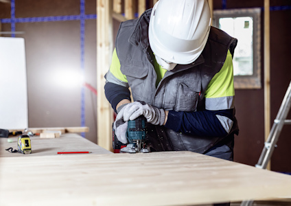 Byggarbetare i hjälm och skyddskläder använder elektriskt verktyg på träskiva inomhus.