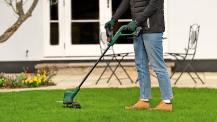 Person använder en grön grästrimmer på en gräsmatta utanför ett hus.