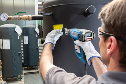 Person använder blå värmepistol på industriell utrustning, bär skyddsglasögon och handskar.