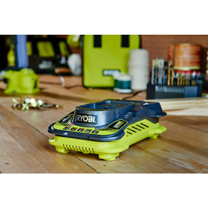 Batteriladdare från Ryobi placerad på ett träbord, omgiven av verktyg och tillbehör i bakgrunden.