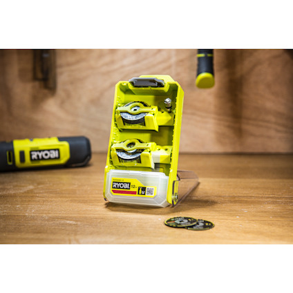 Grön hållare med tre cirkelsågblad från Ryobi, placerad på en träyta. Bakgrund med hängande verktyg.