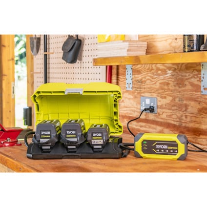 Batteriryggsäck av märket RYOBI med tre batterier och en grön förvaringslåda på en arbetsbänk i en verkstad.
