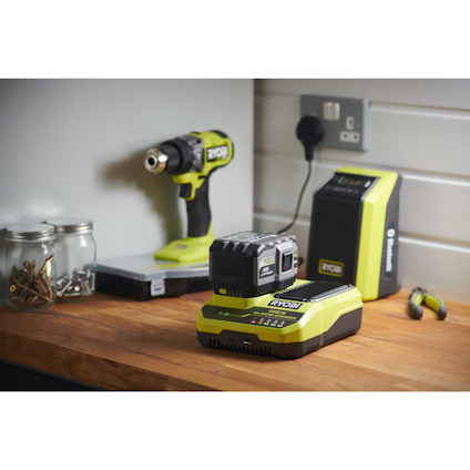 Batteridriven borr, snabbladdare och batteri på träbord med glasburkar och verktyg. Märke: Ryobi