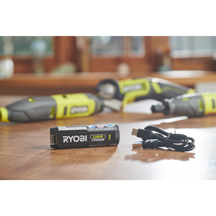Batteri märkt "RYOBI USB Lithium" och en USB-kabel ligger på ett bord, med verktyg i bakgrunden.
