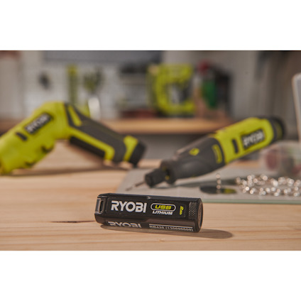 Ryobi USB Lithium-batteri på trämaterial, med oskarpa verktyg i bakgrunden.