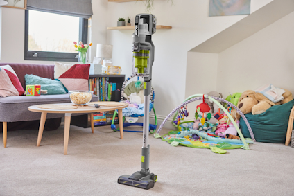 Skaftdammsugare av märket RYOBI på ett ljust vardagsrumsgolv, bredvid en lekmatta och soffbord.