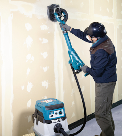 Person använder en slipmaskin på en spacklad vägg, ansluten till en Makita dammsugare.