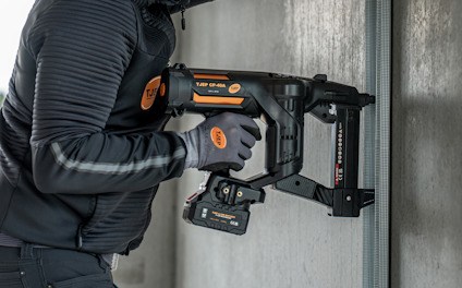 Person håller en spikpistol mot en betongvägg, iförd svarta arbetskläder och handskar.