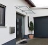 Modernt hus med en glas- och metallbaldakin över ytterdörren, ett grått garage och en krukväxt bredvid ingången.