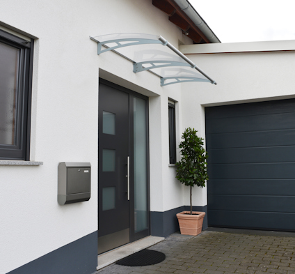 Modernt hus med en glas- och metallbaldakin över ytterdörren, ett grått garage och en krukväxt bredvid ingången.
